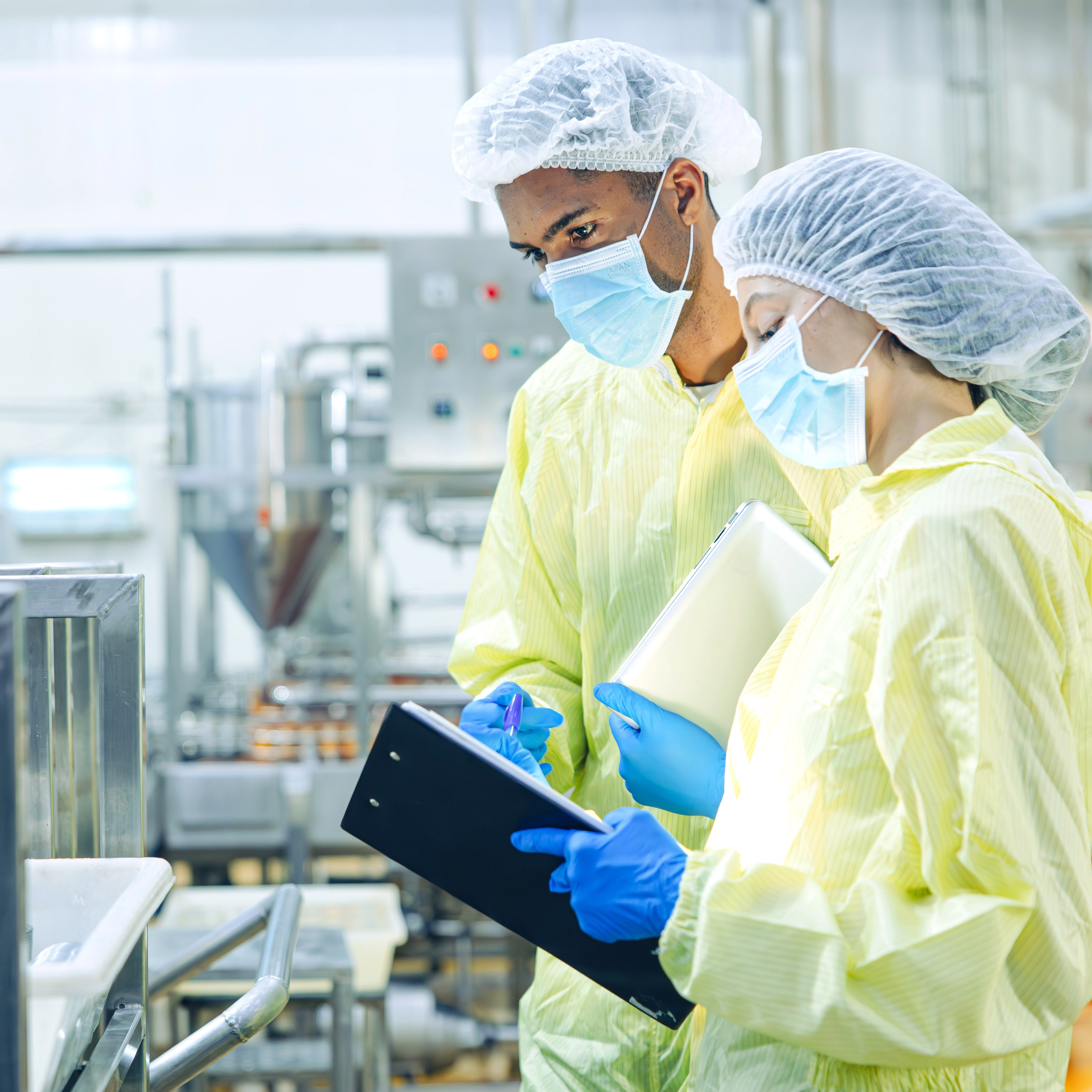  Two figures in quality assurance, dressed in clean room equipment, including hairnets, face masks, and gloves, in a laboratory setting, assuring the safety and efficacy of a process or product in biopharmaceutical and biological production.